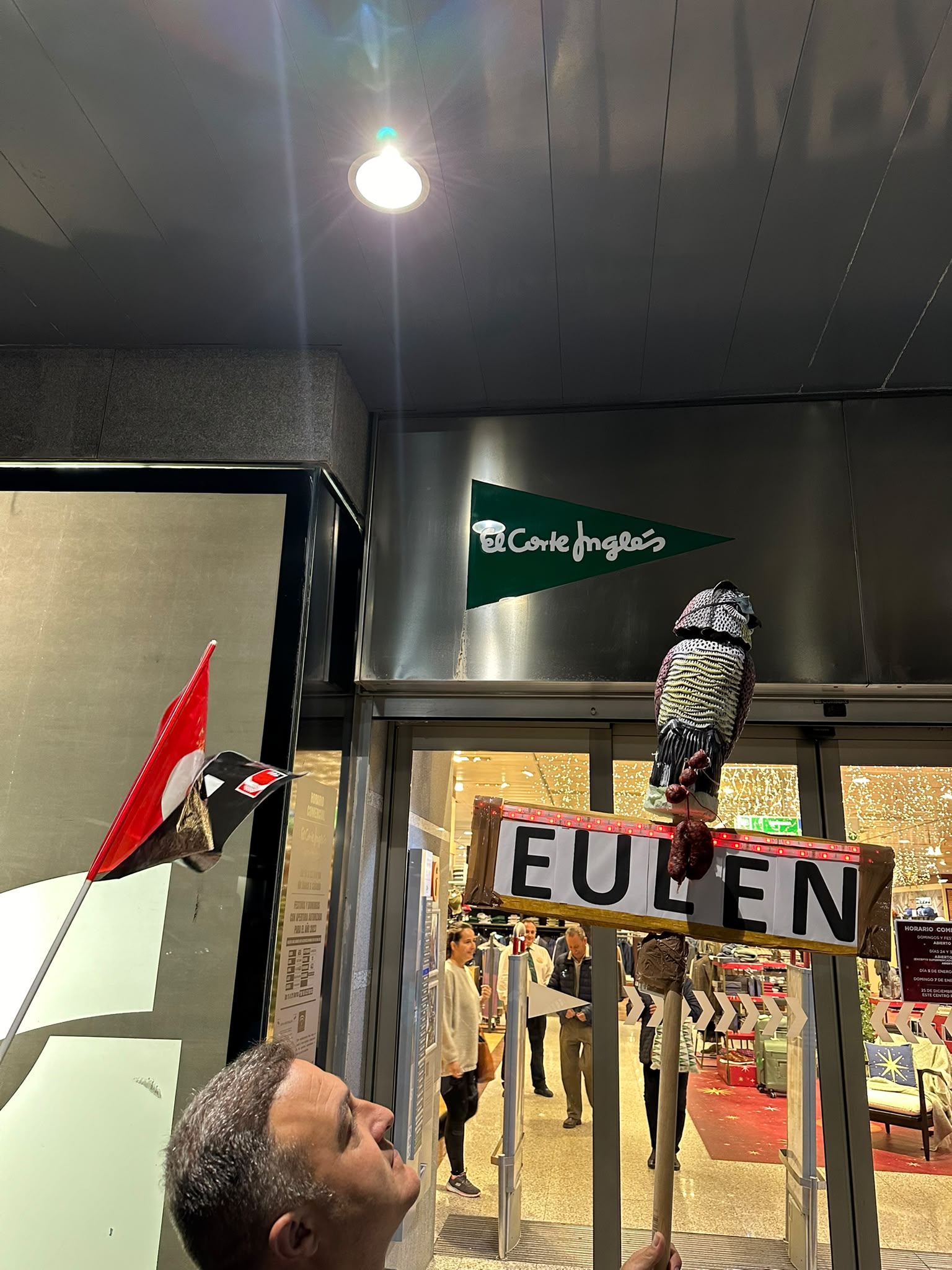 Figura de un mochuelo sobre un cartel de EULEN junto a una bandera de CGT ante la entrada de El Corte Inglés