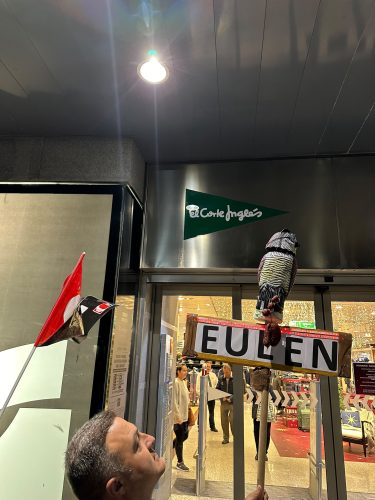 Figura de un mochuelo sobre un cartel de EULEN junto a una bandera de CGT ante la entrada de El Corte Inglés